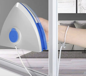 A triangular wall-mounted window air conditioner or fan unit with a white body and blue accents, shown attached to a glass window in an indoor room. A person's arm is visible holding or adjusting it, and a cord hangs from the unit.